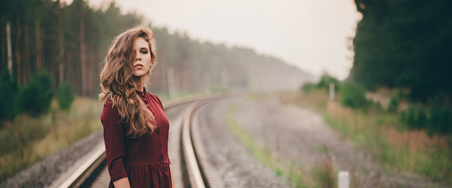 Beautiful girl with curly natural hair enjoy nature in forest on railway. Dreamer lady in burgundy dress walk on railroad. Female portrait of inspired girl on rails at dawn. Sun in hair in autumn.