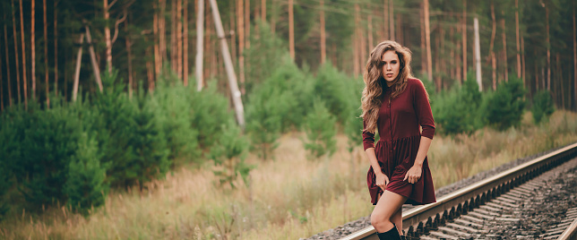 Beautiful dancing girl with curly natural hair enjoy nature in forest on railway. Dreamer lady in burgundy dress walk on railroad. Dance of inspired girl on rails at dawn. Sun in hair in autumn.