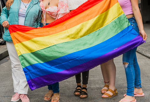 gay or LGBTIQ+ community flag held by a group of women in the background where only their feet are shown.
