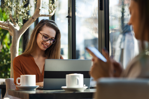 Two young business women in a cafe having one on one meeting. Friends after work talking gossiping and having coffee at a window table on a sunny day.