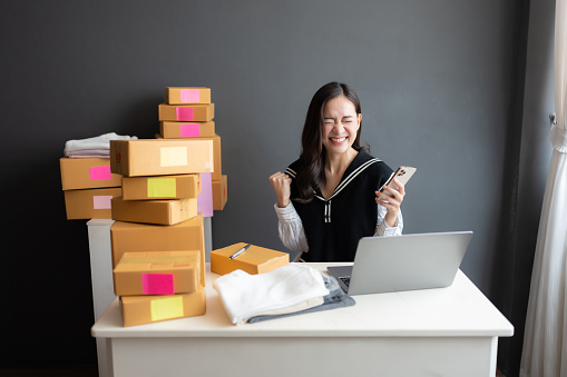 Asian female online shopping business owner excited Due to the target sales volume, holding a phone, having a laptop on desk. There is a customer's product box waiting to be sent to the post office.