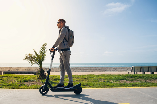 Young man traveler riding e-scooter along the sea coast and beach at sunny summer day. Male driving electric scooter outdoors. Sustainable transport and travel. Sustainable lifestyle concept