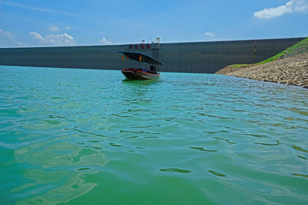 Upstream view on the dam's crest with a long-tailed boat. Surrounded by mountains and lush nature.