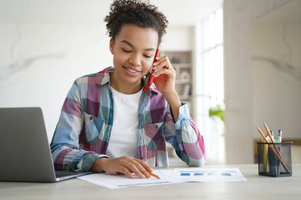 menina adolescente mestiça estudante chamada por telefone, discutindo lição de casa com amigo, sentado na mesa - deside - fotografias e filmes do acervo