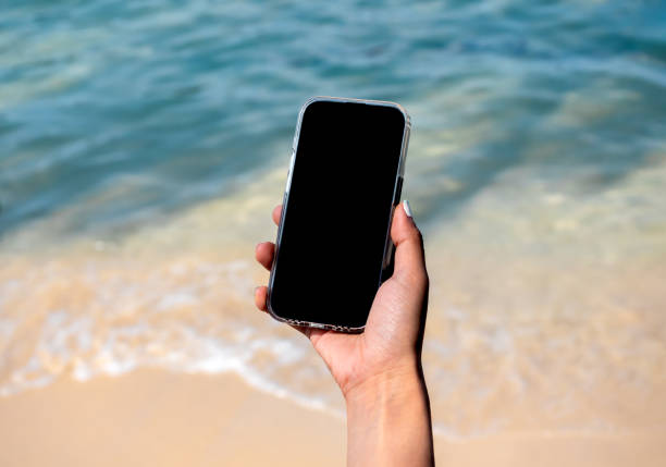 istock Mobile beach phone with empty screen. Mockup image of black blank desktop screen in hand holding mobile smart phone on the blue sea background in summer. Travel booking, searching, business marketing. 1495811135
