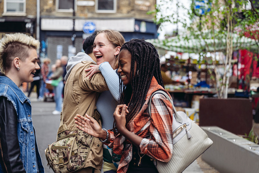 Casually dressed friends in late teens and early 20s laughing and hugging as they meet up for weekend shopping and fun on Brick Lane.