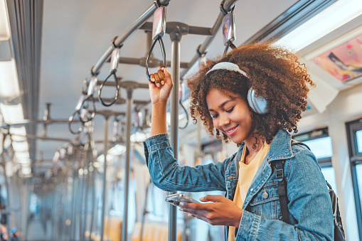 Happy young African American woman passenger listening music via smart mobile phone in a train, Smile female wearing wireless headphones while moving in the tram, lifestyle, transportation.