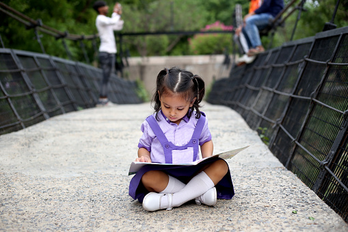 Small girl of schoolgirl sitting in the middle of suspension bridge and studying carefully.