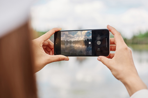 Unrecognizable female blogger using cellular technology for shooting video content during travel vacations, woman with smartphone device photographing lake or river.