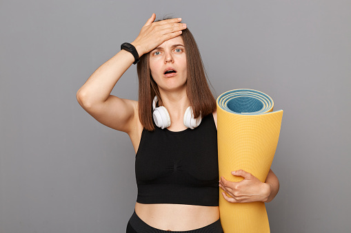 Shocked girl holding fitness mat in her hands posing isolated over gray background forgot about time of training showing facepalm gesture looking at camera with open mouth.