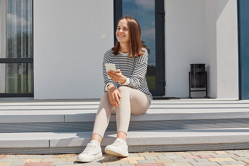 Dark haired young adult woman wearing striped shirt using phone posing near house sitting on porch holding smartphone looking away checking social networks.
