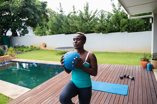 A mid adult African woman doing some exercises in her garden at home. Woman keeping fit and healthy by doing a home workout. Step up's with a weighted medicine ball.
