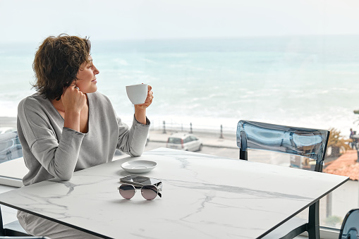 Freelance business woman enjoying a fragrant coffee alone in cafe in the morning near panoramic seaside window.