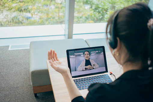 The Asian businesswoman is using a laptop and a headset microphone for video calls, participating in remote meetings to discuss matters with colleagues or work partners.