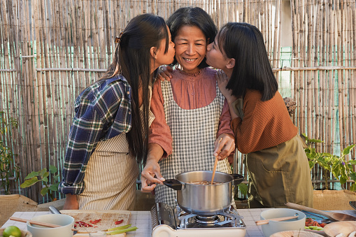 Happy asian mother having tender moment with adult daughters while cooking traditional thai soup at home terrace