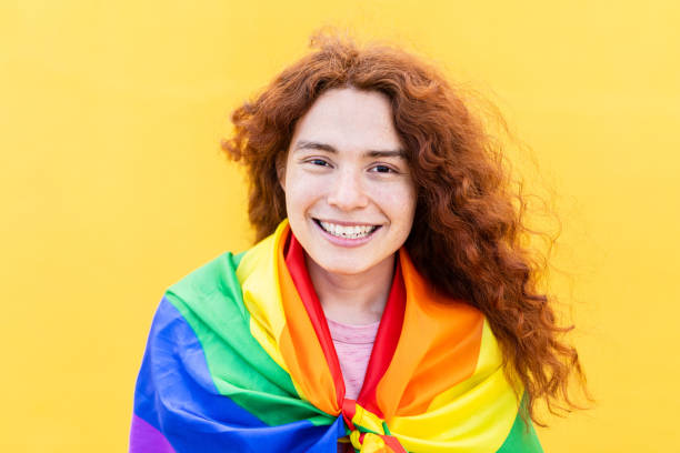 Portrait of non binary gay man with rainbow flag over yellow background stock photo