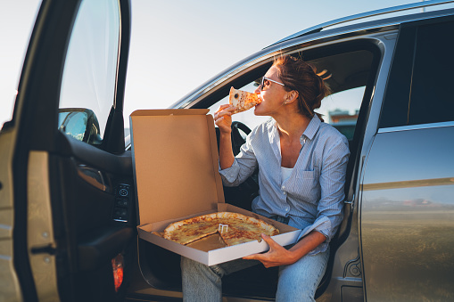 Midle-aged woman eating just cooked italian pizza sitting on driver car seat during meal break and enjoying sunset light. Auto traveling, fast food eating or car jorney lunch break concept image.