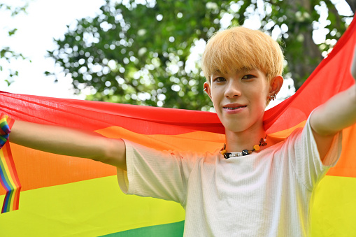 Portrait with LGBT teenage boy with bright colored hair smiling proudly and holding pride flag with outdoor background.