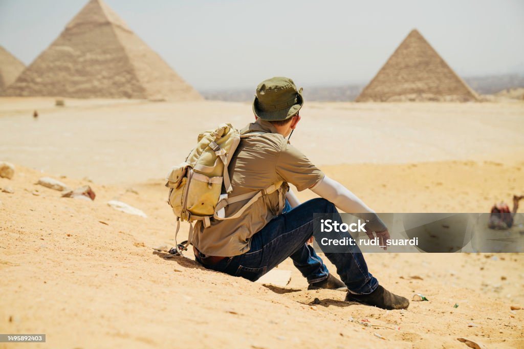 Egypt, Cairo, asian male tourist sitting  on rocks with Great Pyramid of Giza in background Egypt, Cairo, asian man tourist sitting  on rocks with Great Pyramid of Giza in background Desert Area Stock Photo