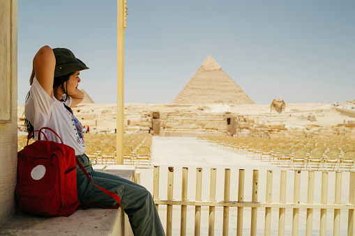 Egypt, Cairo, asian female tourist sitting  on fence with Great Pyramid of Giza in background