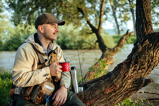 Male fisherman sitting on a tree and enjoying coffee