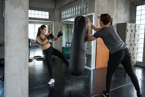 Asian Female boxer kicking sand bag held by personal trainer at fitness gym. Athletic woman practice Thai boxing with coach for bodybuilding and healthy lifestyle concept.