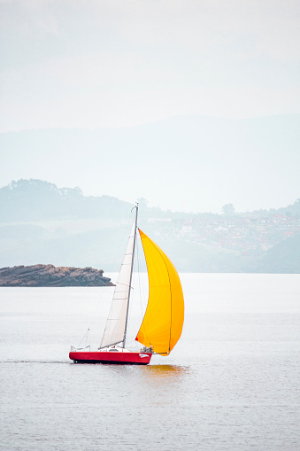 Small sailboat with red hull, sailing the sea with a large yellow sail unfurled, standing out with its colors on the horizon, concepts, vertical shot.