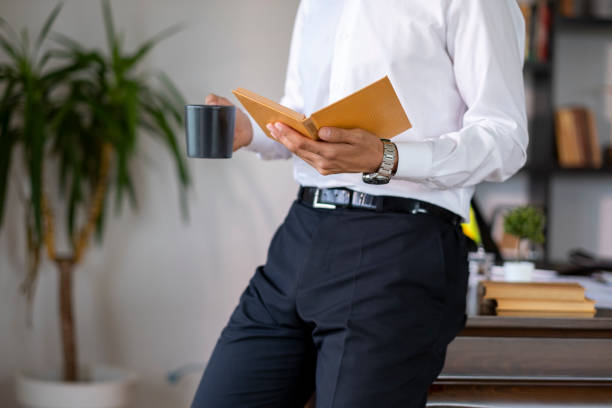 unrecognizable businessman reading a book while drinking coffee in his home office