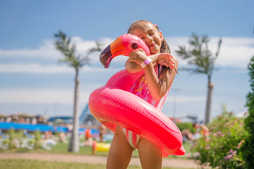A girl in a striped swimsuit hugs a swimming circle in the shape of a flamingo. The child is happy and enjoys his vacation at the resort