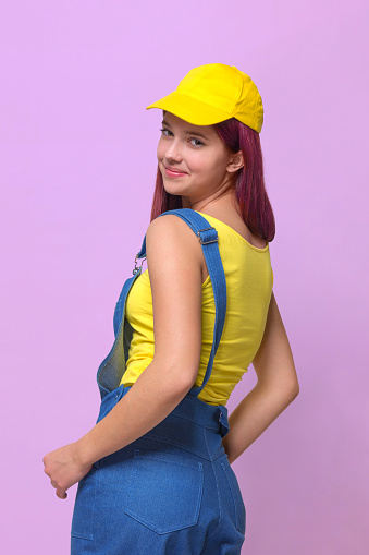 Girl in a blue denim overalls and a yellow T-shirt and a baseball cap on a pink studio background