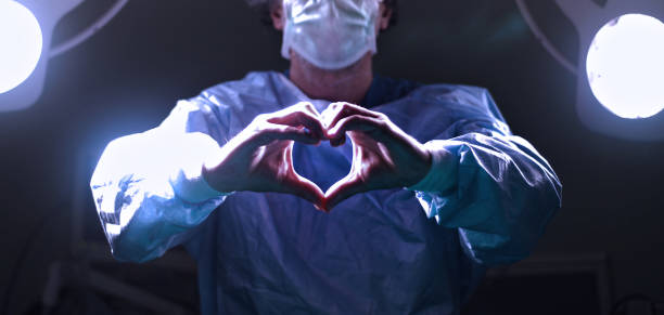 Surgeon making heart symbol and shape with hands in surgery room after successful operation Doctor's blue-green uniform shows a heart hands shape. Medical love, care and safety symbol, health protection sign concept, A medical worker makes a positive symbol with fingers stock photo surgical light stock pictures, royalty-free photos & images