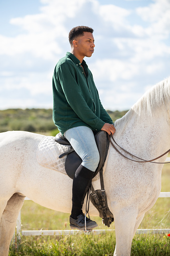 Side view of young African American male looking away while sitting on back of white horse on ranch