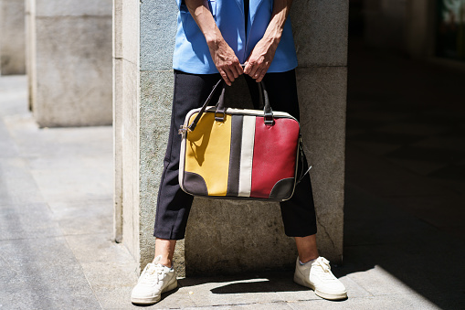 Unrecognizable female in casual outfit carrying colorful handbag standing standing on paved city street in daylight
