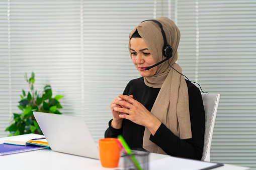 Muslim woman wear headset looks at laptop talk by video call in office