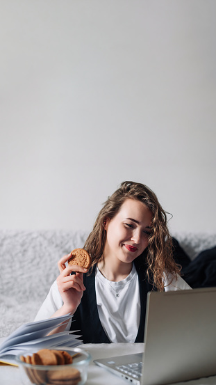 Young woman watching series or podcasts looking at the screen with interest holding cookies in hand sitting at the kitchen table with a glass jar and lapto, smiling happily Unconscious eating disorder