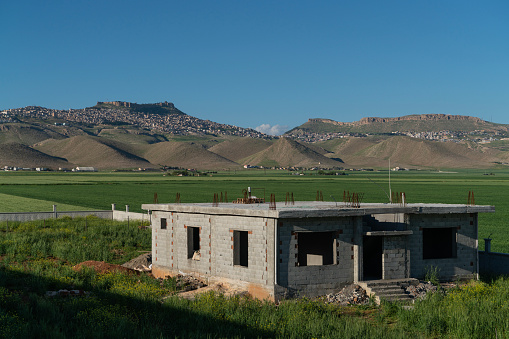 Reinforced concrete construction on the fertile agricultural lands of the Mesopotamian plain, the historical city of Mardin is seen on the summit of the mountain in the background. Reinforced concrete house construction is seen among the wheat fields. Shot with a full-frame camera in daylight.