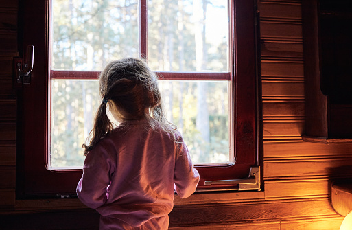 Small girl just woke up in beautiful cabin, standing on the bed in pajama, looking trough the window.