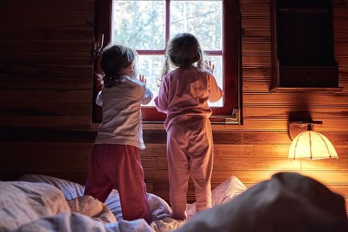 Small girls just woke up in beautiful cabin, standing on the bed in pajamas, looking trough the window.