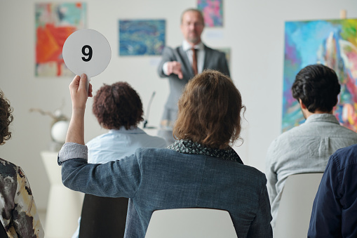 Rear view of man raising his tablet with number up while sitting at auction in art gallery among other people