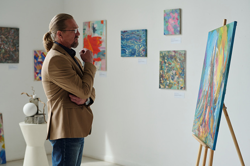 Mature man standing in front of canvas with painting and enjoying the modern art in gallery