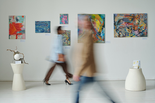 Blurred motion of people visiting art gallery, they walking along the hall among paintings on the wall