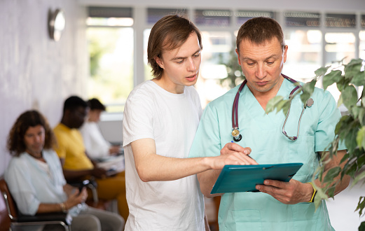 Serious middle-aged male doctor discussing medical document with concerned patient standing in waiting room