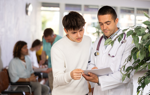 Young male doctor and patient discussing medical document standing in waiting room