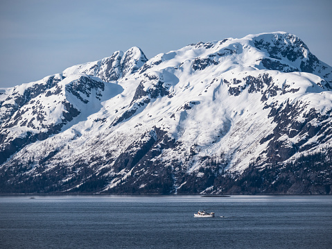 Cruise ship point of view across calm waters toward snowcapped mountain range and small boat detailing scale. Springtime in Southeastern Alaska.