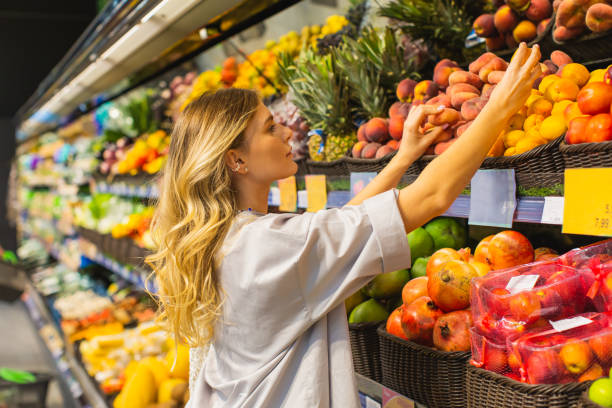 Young blonde woman chooses peach in fruit section. Autumn market. Vegetarianism. Healthy food. Choice. Shopping. Select fruits by scent. Sniff food stock photo