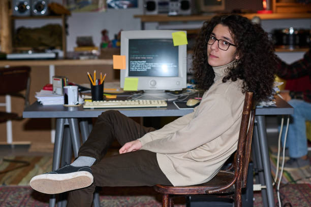 Portrait of young programmer with curly hair looking at camera sitting at his workplace with computer