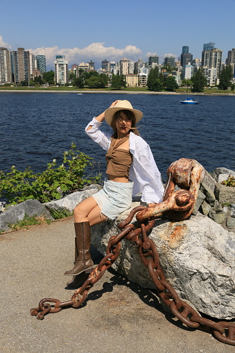 A Japanese model sitting on a rock with a boat anchor on a sunny, windy Spring day with the city of Vancouver BC in the background. She is wearing long blond wavy hair, a hat, brown crop top, white open shirt, blue mini skirt and brown boots.