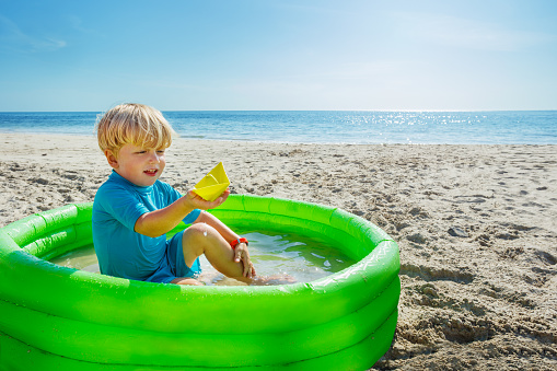 Little happy blond boy play in inflatable pool at the beach holding plastic toy ship