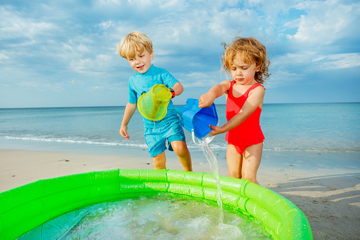 Two happy kids play to inflatable pool bringing water from sea in buckets and splash inside