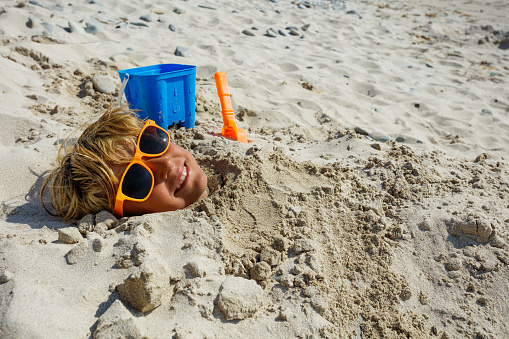 Photo of the boy's face dug into sand with all covered but the head wearing sunglasses, smile, toy bucket and scoop aside